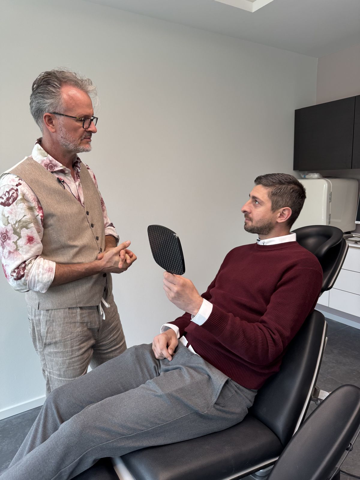 Dr. Twent bespricht mit einem Patienten die Behandlung, der Patient hält einen Handspiegel.